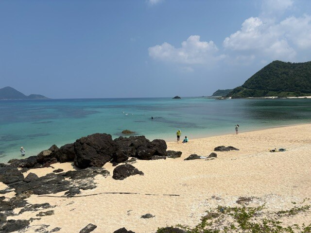 A shoreline of white sand and dark volcanic rock. The shallows are pale emerald, deepening to blue further out, with a green headland jutting into the sea in the distance